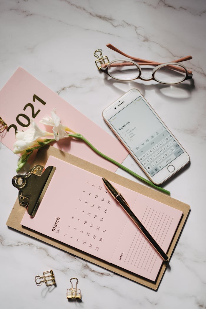 Stylish flat lay of pink stationery on marble, featuring a calendar, smartphone, and eyeglasses.